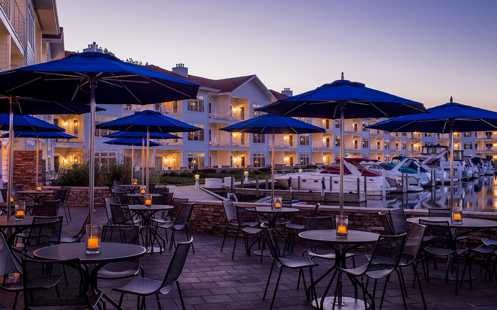 outdoor restaurant patio at twilight with candles, metal tables and chairs, navy blue umbrellas and a view of hotel exterior, marina and boats