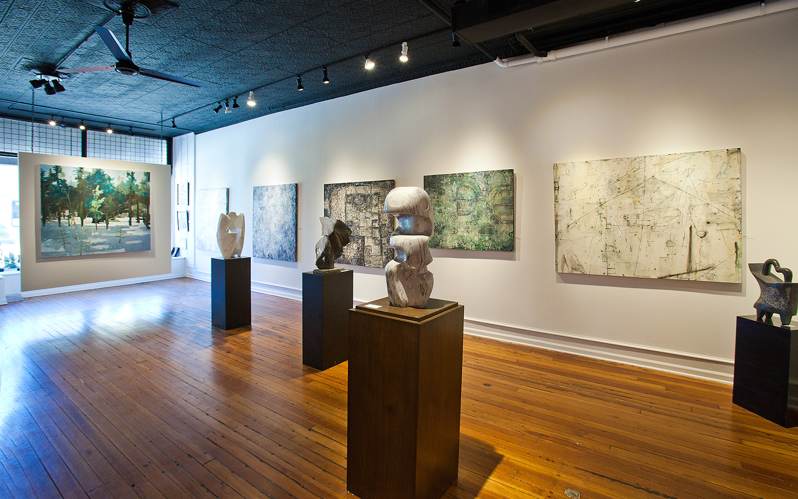 art gallery with sculptures displayed on wood pedestals and abstract green and nuetral-colored paintings adorn the walls
