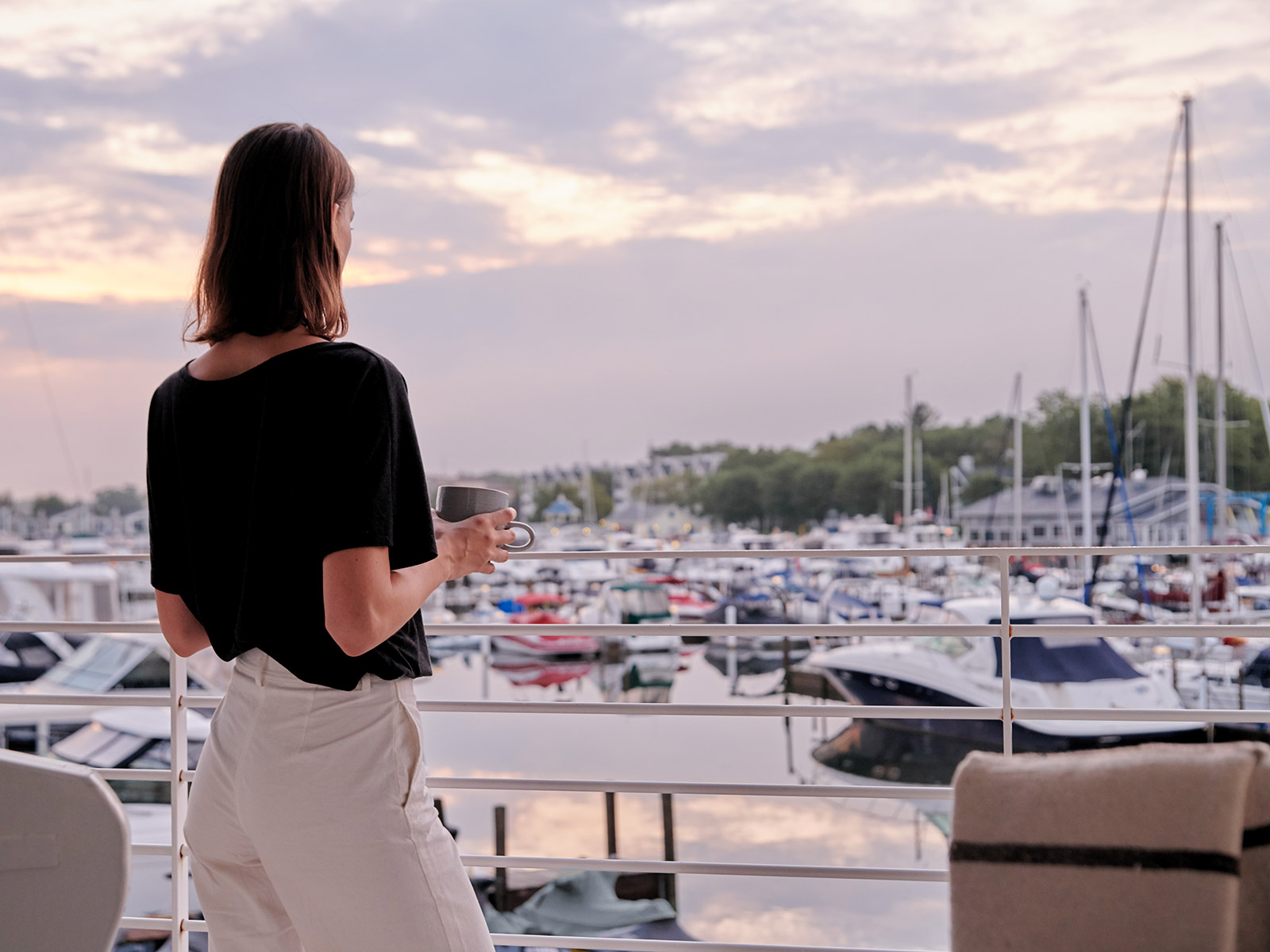 Woman with black shirt and white pants looking out from balcony at pink skies and boats in the New Buffalo Marina