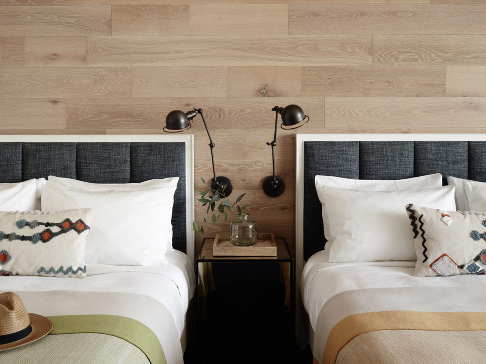 Two queen beds with blue texture headboards, wood wall, black sconces and a woven hat