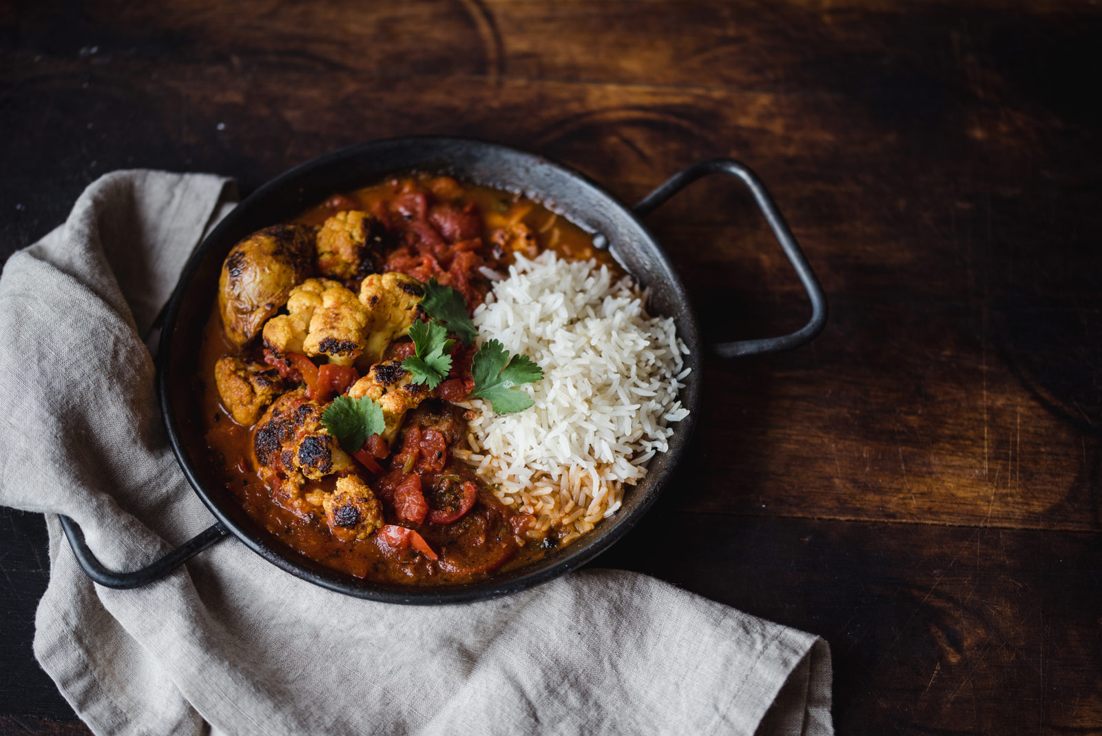 Cauliflower_Tikka Black cast-iron serving dish filled with cauliflower tikka masala and basmati rice, garnished with cilantro and placed on a dark wood table with natural colored cloth napkin on the side