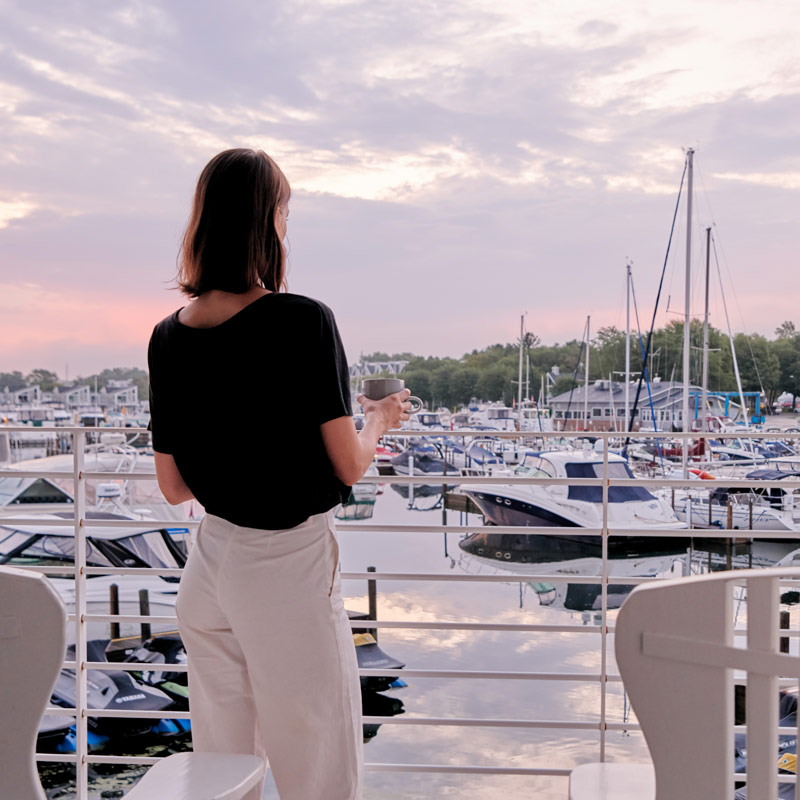 Woman with black shirt and white pants looking out from balcony at pink skies and boats in the New Buffalo Marina