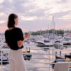 Woman with black shirt and white pants looking out from balcony at pink skies and boats in the New Buffalo Marina