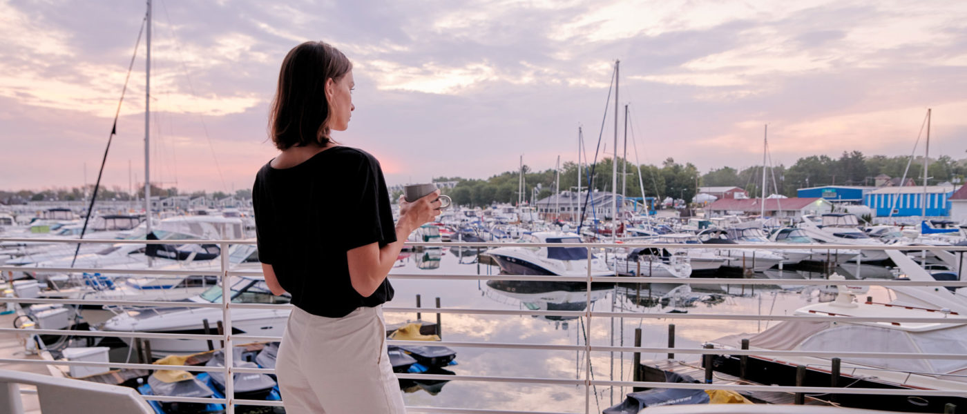 Woman with black shirt and white pants looking out from balcony at pink skies and boats in the New Buffalo Marina