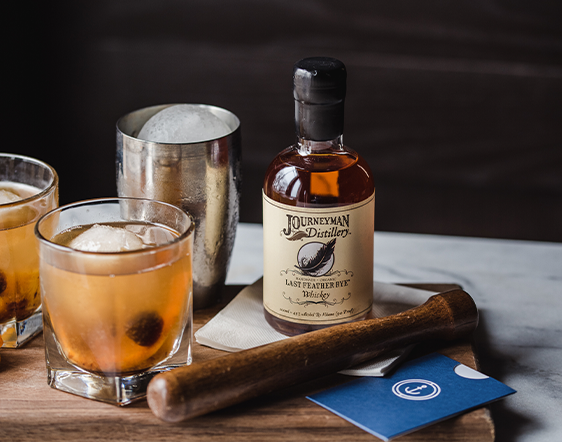 Half pint bottle of Journeyman Distillery Rye Whiskey on a wooden board with wood muddler and classic old-fashioned cocktail in a rocks glass.