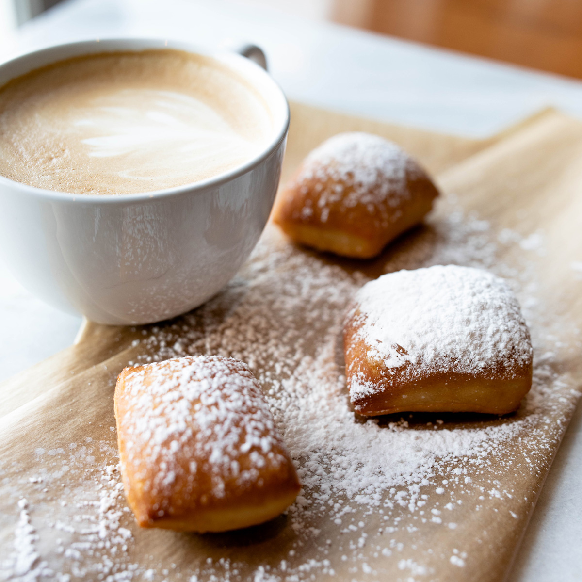 Three fluffy beignets with powdered sugar and a latte in a white mug on a tabletop