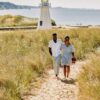 A couple walks through the dune grass with the New Buffalo lighthouse in the background.