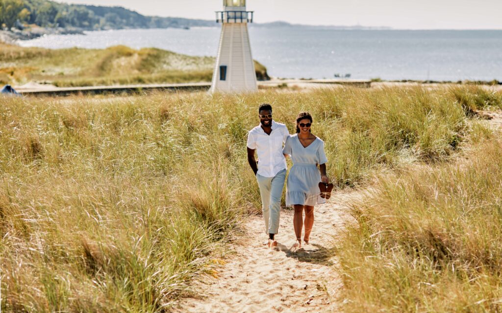 A couple walks through the dune grass with the New Buffalo lighthouse in the background.