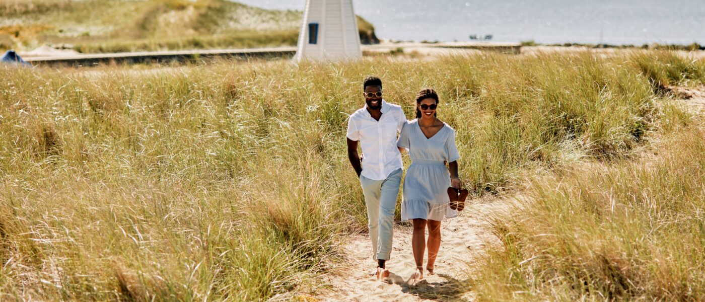 A couple walks through the dune grass with the New Buffalo lighthouse in the background.